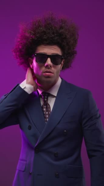 sexy elegante hombre de negocios en traje mirando a un lado, sosteniendo la mano detrás del cuello, la fijación de gafas de sol y mirando hacia abajo en frente de fondo colorido - Metraje, vídeo