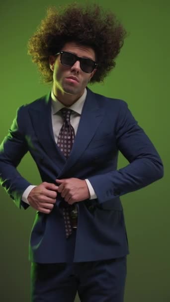 fresco hombre de negocios elegante con gafas de sol cogidas de la mano en los bolsillos, abotonando traje y mirando a un lado sobre fondo colorido - Metraje, vídeo