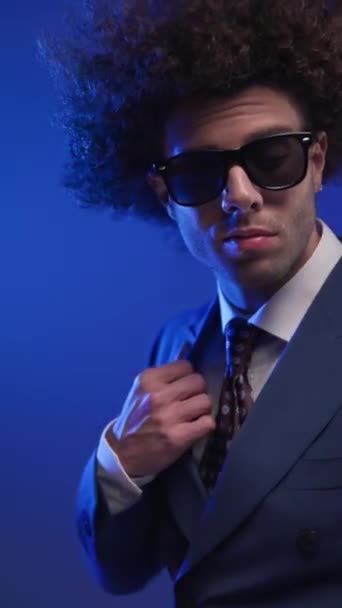 Nahaufnahme-Video von sexy lockigem Haar Mann mit Sonnenbrille Anordnung Anzug beim Blick hinter, berühren Hals und selbstbewusst vor buntem Hintergrund - Filmmaterial, Video