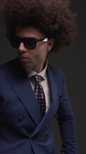 sexy hombre de negocios elegante con gafas mirando a un lado, la organización de traje, sonriendo y sosteniendo la mano en el bolsillo, tocando templo y posando frente a las luces de flash sobre fondo gris - Imágenes, Vídeo