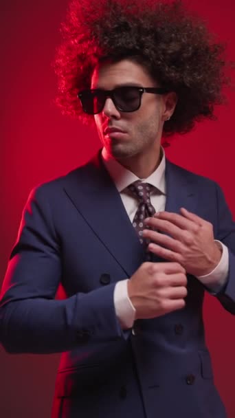 pionowy film seksownego młodego mężczyzny pocierającego dłonie, patrzącego w bok, dopasowującego garnitur i robota poruszającego się przed czerwonym tłem - Materiał filmowy, wideo
