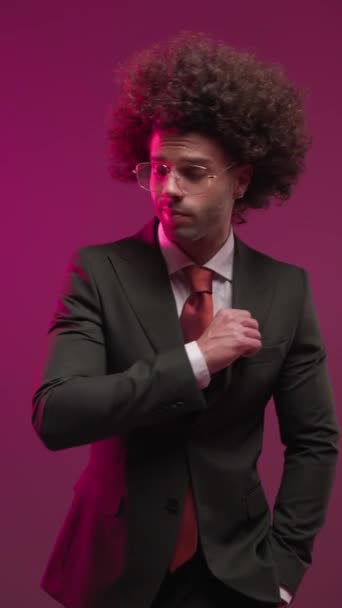 pohledný mladý podnikatel s brýlemi rozhlížející se na stranu, držící ruku v kapse, upravující černý oblek a tření dlaní před barevným pozadím - Záběry, video