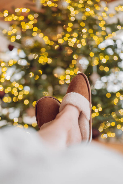 Vacances de Noël et Nouvel An. Jambes en daim brun pantoufles de fourrure sur un arbre de Noël dans des guirlandes fond. Confort et vacances d'hiver. humeur confortable. - Photo, image