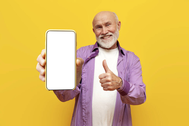 old bald grandfather shows like and blank smartphone screen on yellow isolated background, pensioner with phone recommends and advertises phone display online - Photo, Image