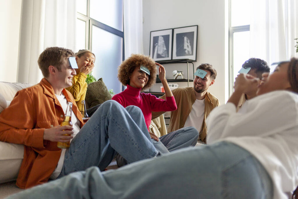 πολυφυλετική ομάδα νεαρών φίλων μαθητών που παίζουν παντομίμα μαζί και πίνουν μπύρα στο πάρτι στο σπίτι, νέοι που κάθονται στο πάτωμα με ποπ κορν και μαντεύουν το χαρακτήρα από το παιχνίδι - Φωτογραφία, εικόνα