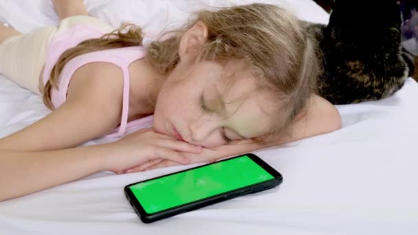 Egy gyerek egy zöld képernyős telefon és egy fekete macska mellett alszik. Krómozott zöld vászon. Kommunikáció, játék vagy rajzfilm a telefonon. - Felvétel, videó