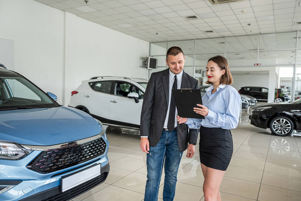 Blanke vrouwelijke verkoper geeft advies aan mannelijke klant in moderne mooie auto showroom. Een man kiest een nieuwe auto bij een autodealer. Het concept van de verkoop en aankoop van auto 's. - Foto, afbeelding
