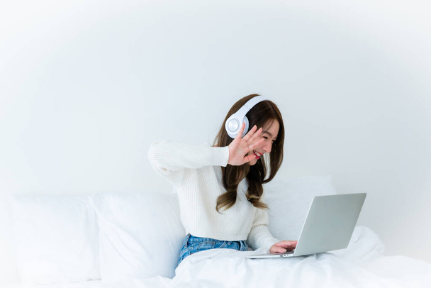 Mattina tempo in camera da letto giovane asiatico donna felice lavoro su computer portatile, indossando auricolare, seduto sul letto a casa - Foto, immagini