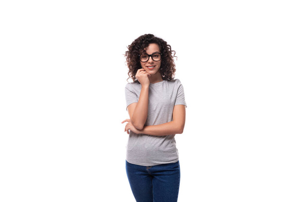 молодая симпатичная брюнетка кудрявая кавказка в очках в джинсах и футболке с распечатанным макетом на белом фоне. - Фото, изображение