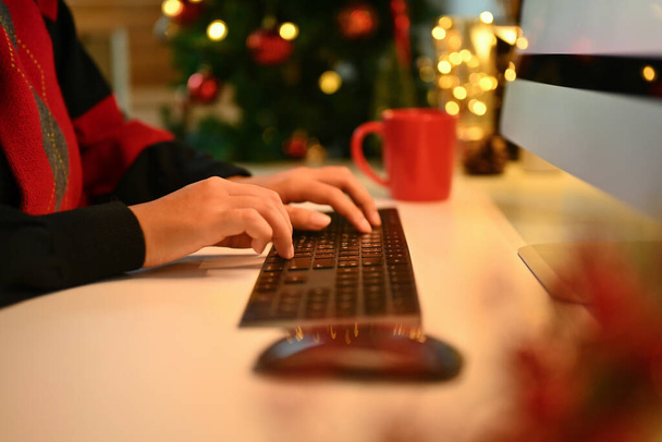 Closeup woman hands typing on computer keyboard against Christmas tree with lights on background. - Photo, Image