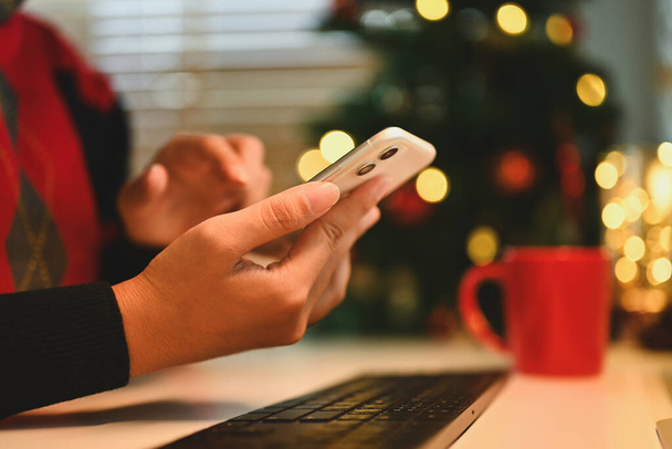Tiro cortado de mulher jovem usando telefone celular perto da árvore de Natal em casa aconchegante. - Foto, Imagem