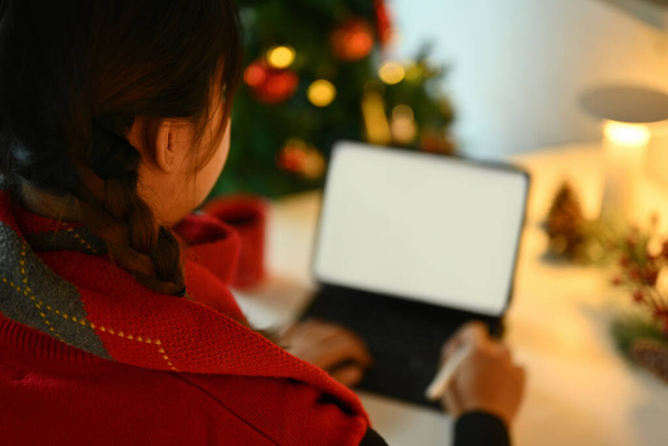 Über-die-Schulter-Ansicht einer jungen Frau im roten Pullover mit digitalem Tablet in der Nähe des geschmückten Weihnachtsbaums. - Foto, Bild