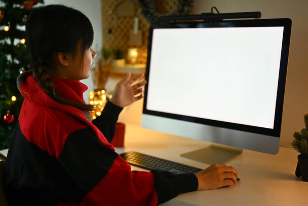 Вид сбоку на молодую женщину в теплом свитере, которая ведет видеозвонок через компьютер, сидя у украшенной елки. - Фото, изображение