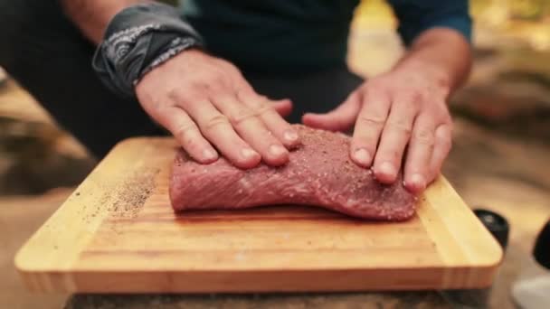 Крупним планом чоловічі руки втирають спеції в м'ясо, готуючи його для приготування їжі на вогні в дикій природі. Маринування м'яса для подальшого приготування на вогні. - Кадри, відео