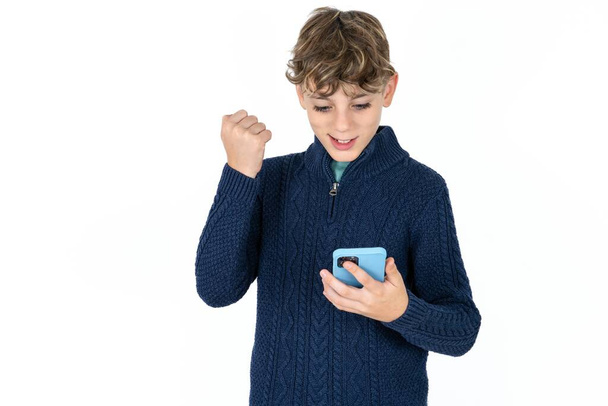 Schöner kaukasischer Teenie-Junge in blauem Pullover, der in der Zelle die Hände hält und aufgeregt die Faust hebt, nachdem er gute Nachrichten gelesen hat. - Foto, Bild