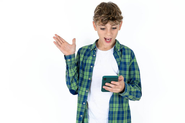 guapo adolescente chico usando camisa a cuadros sobre fondo blanco sosteniendo en las manos celda leer noticias de navegación - Foto, Imagen