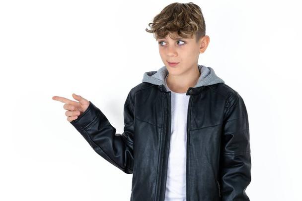 красивий хлопчик-підліток у шкіряній куртці позує на білому фоні студії в копіювальному просторі і щось рекламує, поради кращі ціни. - Фото, зображення