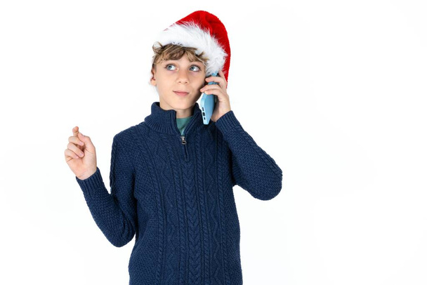 handsome caucasian teen boy in blue sweater and christmas hat speaks on mobile phone spends free time indoors calls to friend. - Photo, Image