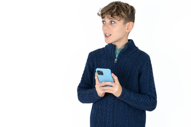 beau garçon adolescent caucasien sur fond de studio blanc tenant un smartphone et regardant de côté l'espace de copie vierge. - Photo, image