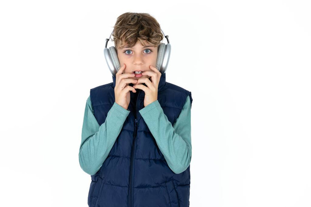 Shocked Handsome Caucasian teen boy in blue vest stares fearful at camera keeps mouth widely opened wears wireless stereo headphones on ears - Valokuva, kuva