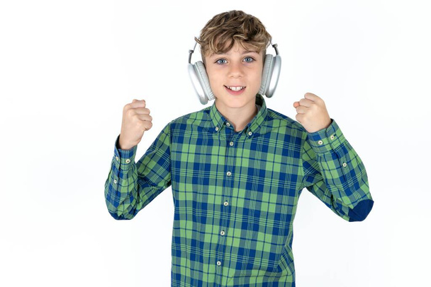 Emocionální pohledný teen chlapec nosí kostkované tričko přes bílé pozadí křičí hlasitě pocit, že vítěz zvedá zaťaté pěsti udržuje ústa otevřená nosí stereo sluchátka na uších dělá ano gesto, poslouchá oblíbené hudby - Fotografie, Obrázek