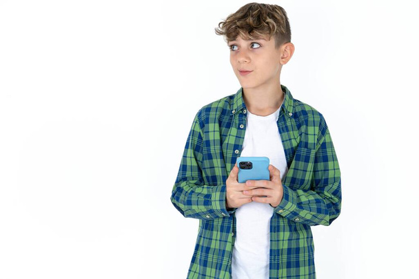 guapo caucásico adolescente chico en blanco estudio fondo sostiene teléfono manos lee buena juventud noticias mirada vacío espacio anuncio - Foto, Imagen