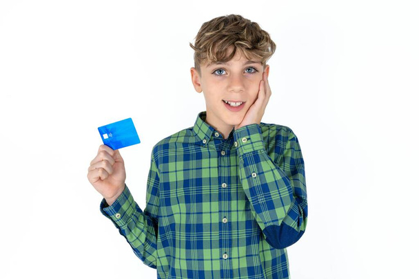 Astonished positive handsome teen boy wearing plaid shirt over white background with arm face hold bank plastic card impressed functions - Foto, imagen