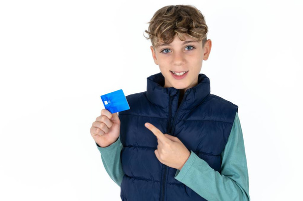 Foto portret van Handsome Kaukasische tiener jongen in blauw vest doet aankoop met wijzende vinger credit bank kaart - Foto, afbeelding