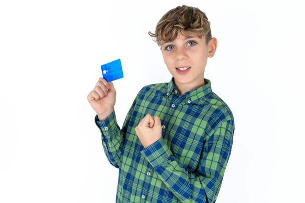 Щасливий позитивний веселий усміхнений гарний підліток у сорочці на білому тлі тримає кредитну картку підняти кулак у перемозі - Фото, зображення