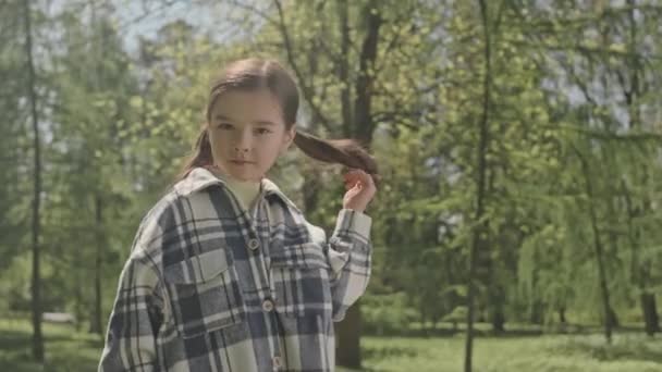 Retrato de niña linda en camisa a cuadros girando el pelo y posando en la cámara en el parque en el día de verano - Imágenes, Vídeo