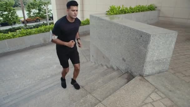 Weitschuss eines Sportlers im schwarzen Outfit mit Armbandhandyhalter, der während des Marathontrainings die Treppe hochläuft - Filmmaterial, Video
