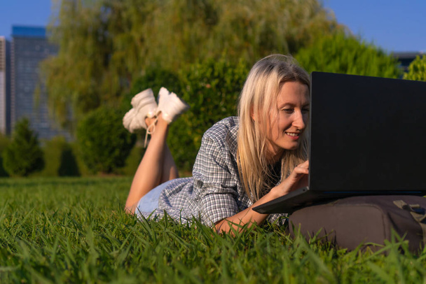 Portret van een jonge glimlachende blonde vrouw op haar buik liggend op het gras in het park met een laptop op een zonnige dag. Studenten communiceren, studeren met een laptop in een stadspark - Foto, afbeelding