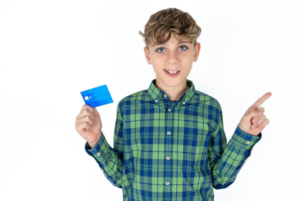 Smiling handsome caucasian teenager boy on white studio background showing debit card pointing finger empty space - Fotoğraf, Görsel