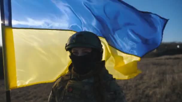 Médico militar femenino del ejército ucraniano mira a la cámara contra ondear bandera azul-amarilla. Retrato de niña en uniforme de camuflaje y casco con bandera de Ucrania en el campo en el día soleado. Dolly disparó - Metraje, vídeo