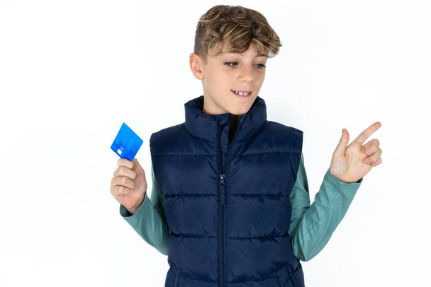 Curioso sonriente guapo caucásico adolescente chico en blanco estudio fondo mostrando banco de plástico mostrando dedo copyspace - Foto, imagen