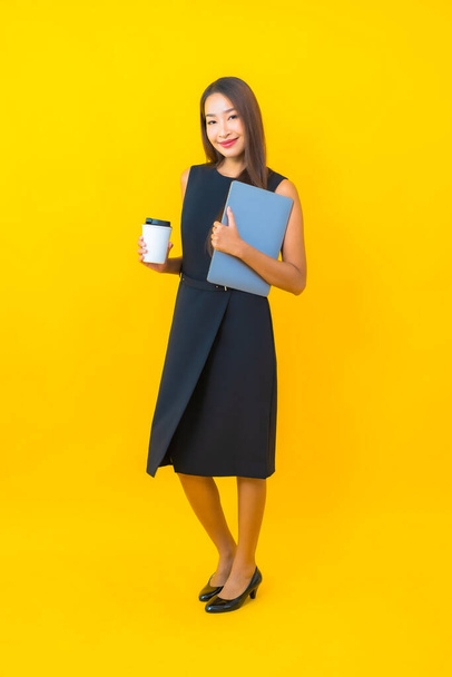 黄色の背景にラップトップコンピュータを持つ肖像画の美しい若いアジアのビジネス女性 - 写真・画像