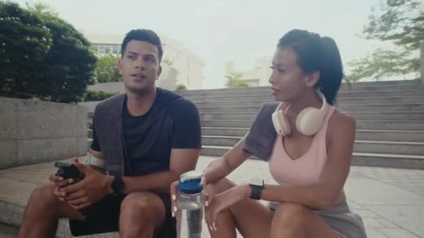 Mediana oportunidad larga de pareja multicultural sentada en los escalones y bebiendo agua mientras descansa después del entrenamiento al aire libre - Imágenes, Vídeo