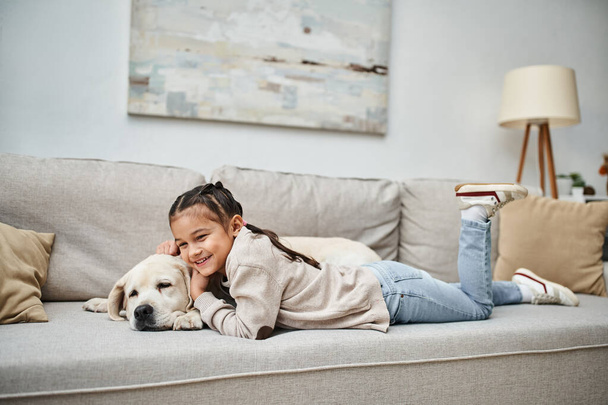 χαρούμενο κορίτσι χαμογελώντας και ξαπλωμένη στον καναπέ με χαριτωμένο λαμπραντόρ στο σύγχρονο σαλόνι, κατοικίδιο ζώο και παιδί - Φωτογραφία, εικόνα