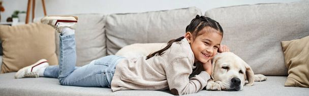 gelukkig meisje glimlachen en liggen op de bank met schattige labrador in moderne woonkamer, huisdier en kind - Foto, afbeelding
