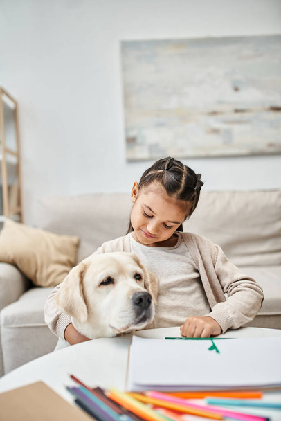 Kleines Mädchen in lässiger Kleidung zeichnet und umarmt Labrador im modernen Wohnzimmer, Kunst und Freizeit - Foto, Bild