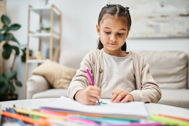 портрет милой девочки начального возраста с цветным карандашом на бумаге в современной квартире - Фото, изображение
