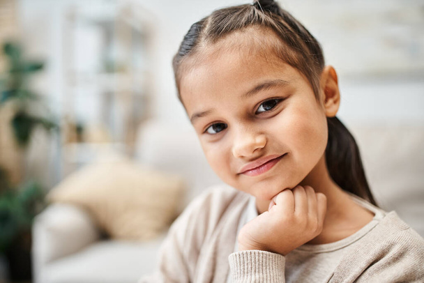 портрет улыбающейся девочки младшего возраста с брюнетками, смотрящей в камеру в современных апартаментах - Фото, изображение