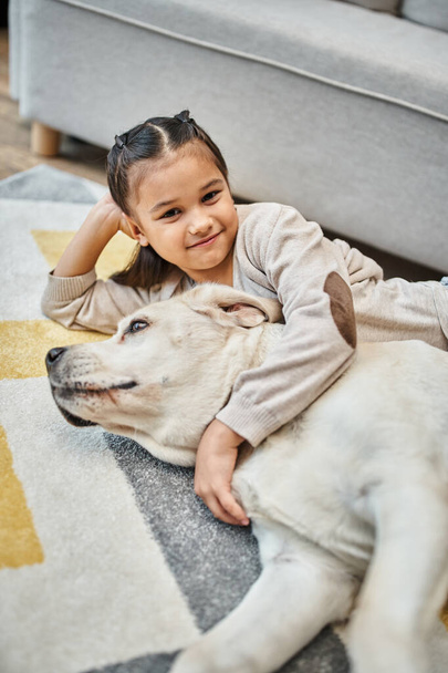 Positives Mädchen in lässiger Kleidung, lächelnder und streichelnder Hund im modernen Wohnzimmer, Kind und Labrador - Foto, Bild
