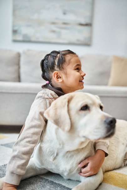 niño alegre en traje casual sonriendo y acariciando perro en la sala de estar moderna, niño y labrador - Foto, Imagen