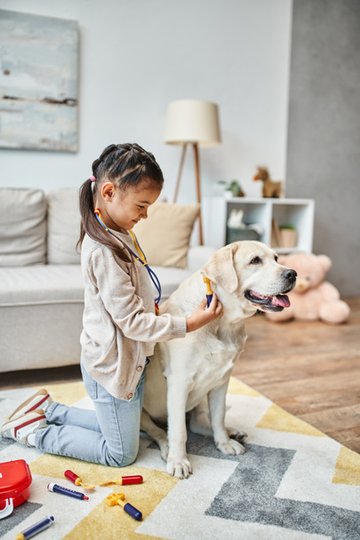 glückliches Mädchen in lässiger Kleidung spielt Arzt mit Labrador im modernen Wohnzimmer, Spielzeug-Verbandskasten - Foto, Bild