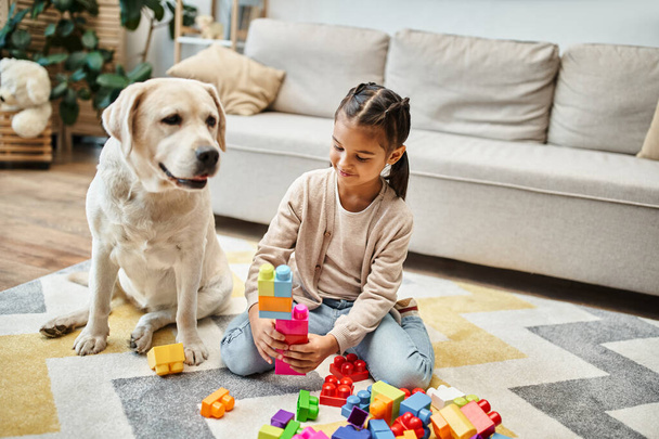 χαμογελαστό κορίτσι παίζει με πολύχρωμα μπλοκ παιχνιδιών κοντά λαμπραντόρ στο σαλόνι, κτίριο πύργος παιχνίδι - Φωτογραφία, εικόνα