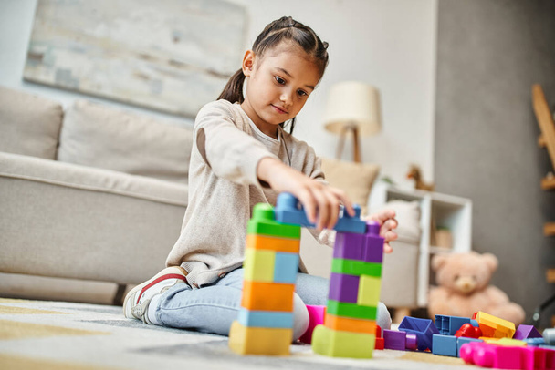 χαριτωμένο κορίτσι παίζει με πολύχρωμα μπλοκ παιχνιδιών στο χαλί στο σύγχρονο σαλόνι, κτίριο πύργο παιχνίδι - Φωτογραφία, εικόνα