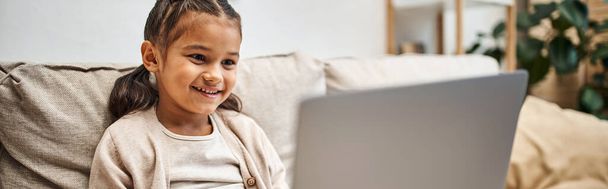 счастливая девушка начального возраста сидит на диване и с помощью ноутбука в современной гостиной, e-learning баннер - Фото, изображение