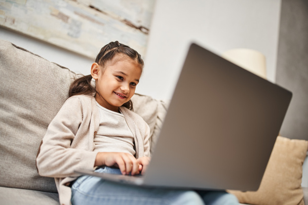 joyful elementary age girl sitting on sofa and using laptop in modern living room, e-learning - Photo, Image