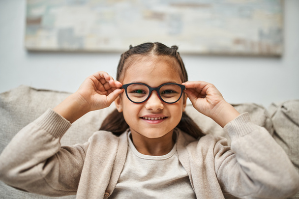 glückliches Mädchen im Grundschulalter in lässiger Kleidung mit Brille und Blick in die Kamera im Wohnzimmer - Foto, Bild